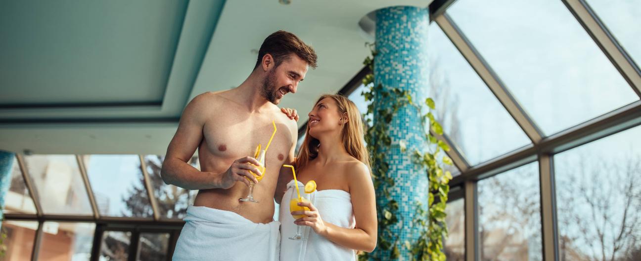 Ein Pärchen genießt einen Cocktail, während beide ein Handtuch tragen. Im Hintergrund sieht man einen Spa-Bereich