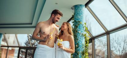 Ein Pärchen genießt einen Cocktail, während beide ein Handtuch tragen. Im Hintergrund sieht man einen Spa-Bereich